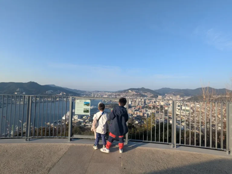 Luca and Nico standing side by side at Mt. Nabekammuri observatory in Nagasaki, quietly overlooking the city as the afternoon sun casts a golden glow across the harbor and skyline, a peaceful moment of reflection at the end of their family adventure.