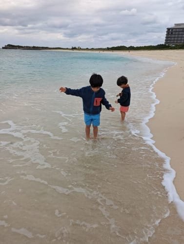 Luca & Nico splashing in the clear waters of Yonaha Maehama Beach, Miyakojima, on a cool day, discovering shells and enjoying family-friendly beach adventure on a Miyako Island Itinerary.