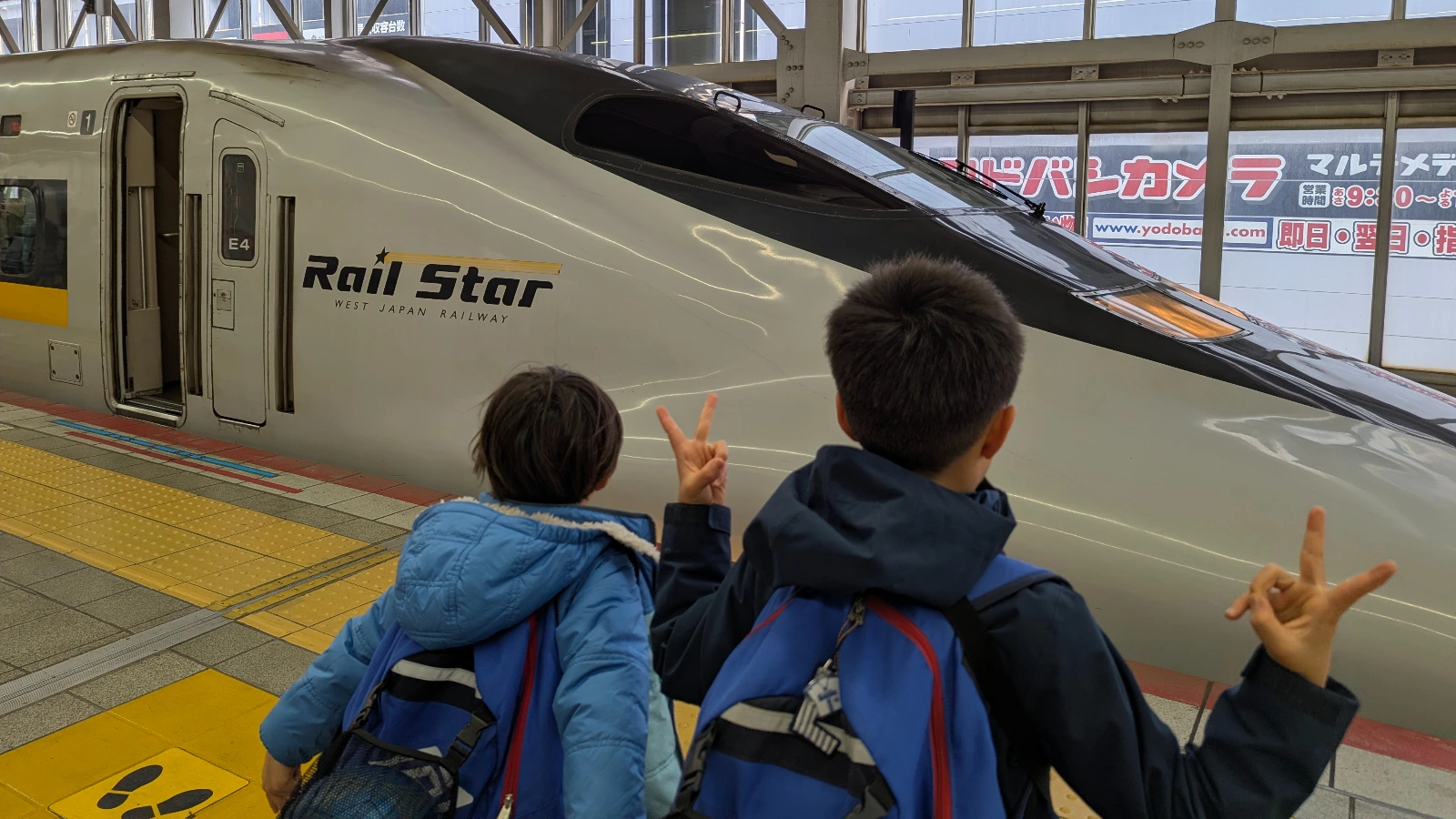 Two children with backpacks posing on a station platform in Japan in front of a Rail Star Shinkansen bullet train, capturing a family train travel moment.
