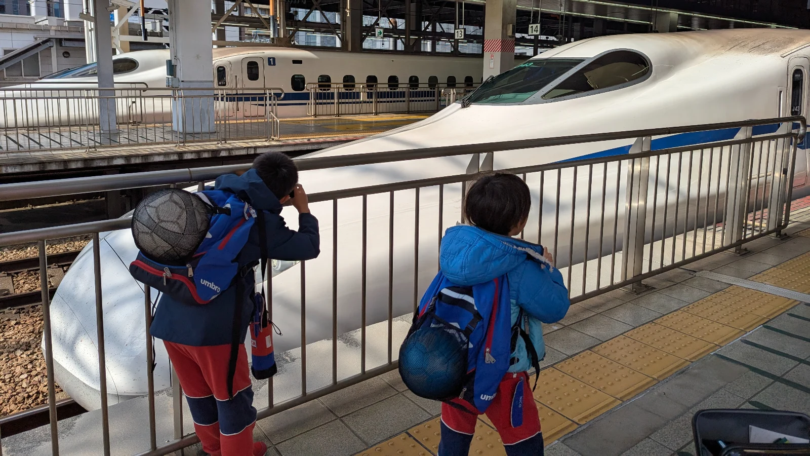 Two children with backpacks watching a Shinkansen bullet train at a Japanese train station platform during a family trip in Japan.