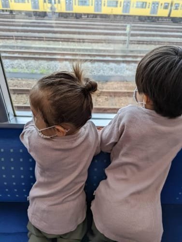 Two siblings sitting on a train in Japan, looking out at a yellow train on adjacent tracks, capturing a moment of fascination with Japan's vibrant train system.