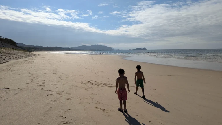 Two children walk barefoot along a wide sandy beach, in Japan, toward the water, sunlight reflecting on the sea and mountains in the distance.