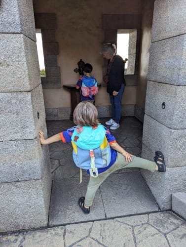 A charming moment at Tokyo DisneySea where two children and their grama explore a stone alcove. One child playfully stretches against the wall while the other inspects a small map, set within a rustic stone structure that reflects the park's nautical and adventure-themed design.