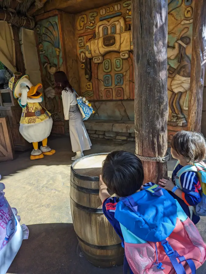 Luca and Nico in colorful Patagonia backpacks peek behind a wooden barrel while watching Donald Duck dressed as an explorer, with vibrant Mayan-themed wall art in the background at Tokyo DisneySea.