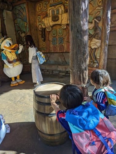 Luca and Nico in colorful Patagonia backpacks peek behind a wooden barrel while watching Donald Duck dressed as an explorer, with vibrant Mayan-themed wall art in the background at Tokyo DisneySea.