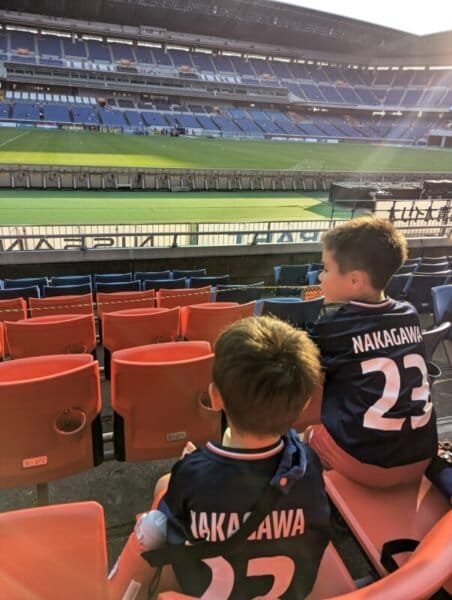 Luca and Nico wearing Yokohama Marinos jerseys sit in the stands of a soccer stadium in Yokohama, Japan, watching a match under the warm evening sunlight.