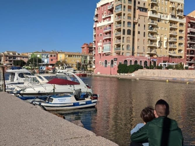 A picturesque vie of Port Saplaya in Valencia, with colorful Mediterranean-style buildings, docked boats, and Papa and Luca by the water, enjoying the serene atmosphere.