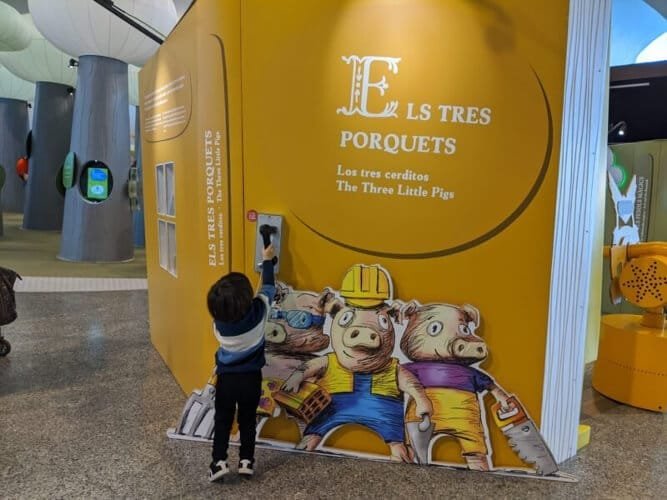 Luca interacting with a colorful educational display about 'The Three Little Pigs' at the Science Museum in Valencia, featuring bright yellow walls and playful illustrations.