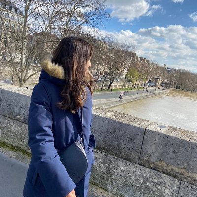 Mama wearing a navy blue winter coat with a fur-lined hood walks along a historic stone bridge overlooking the Seine river in Paris. She gazes at the riverside promenade, with a backdrop of classic Parisian buildings, bare winter trees, and a partly cloudy sky.