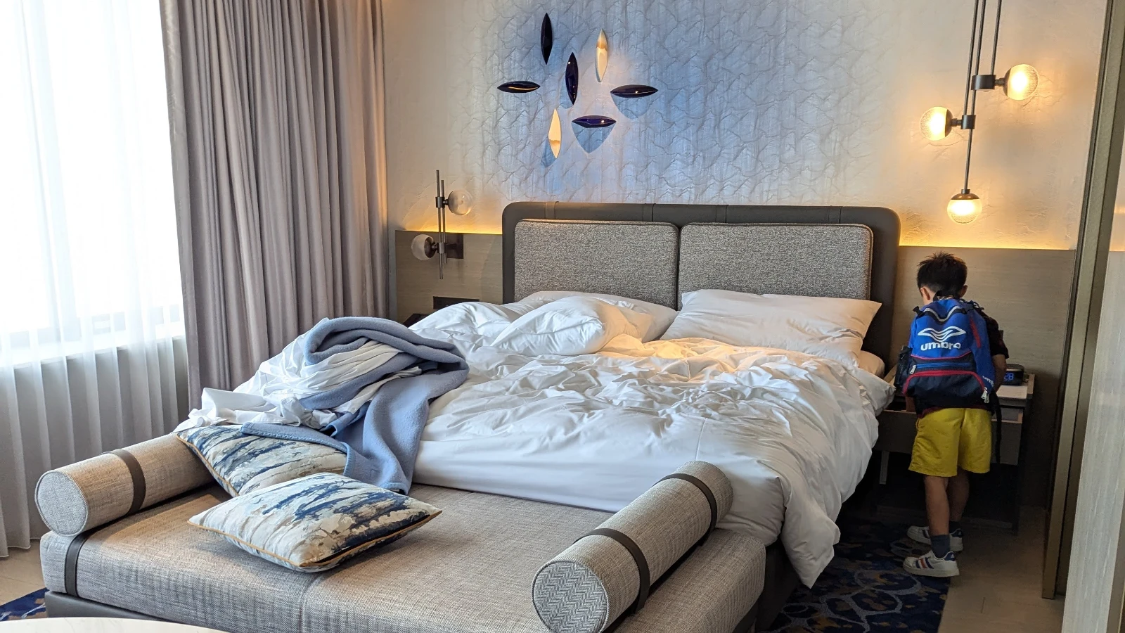Luca with a backpack stands beside the bed in a family room at Marriott Nagasaki, with soft lighting, modern decor, and cozy bedding.