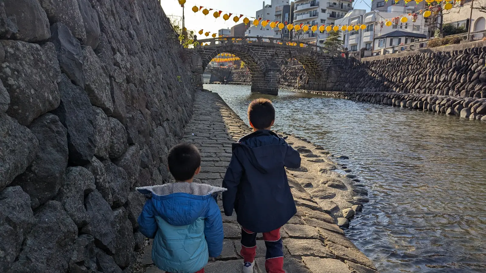 Luca & Nico walk along the riverbank toward Megane Bridge Nagasaki, also known as Spectacles Bridge, during a family adventure in Japan.