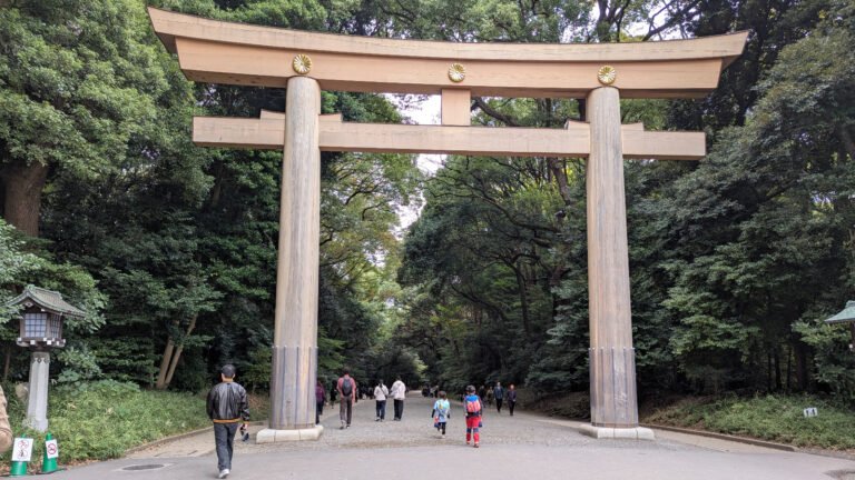Visitors walking under the massive wooden torii gate at Meiji Jingu Shrine in Tokyo, with two children entering along the forested gravel path.