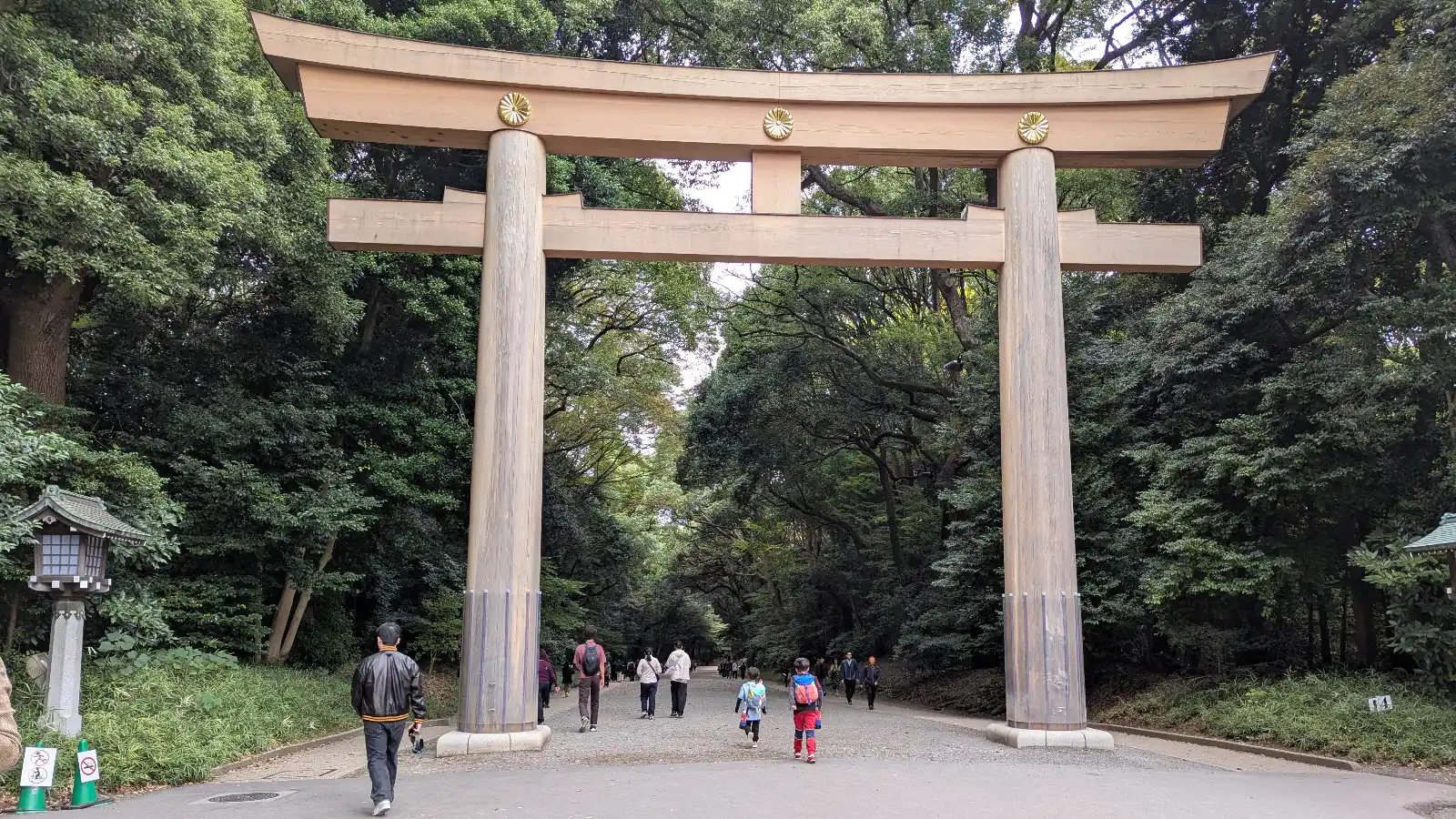 Visitors walking under the massive wooden torii gate at Meiji Jingu Shrine in Tokyo, with two children entering along the forested gravel path.