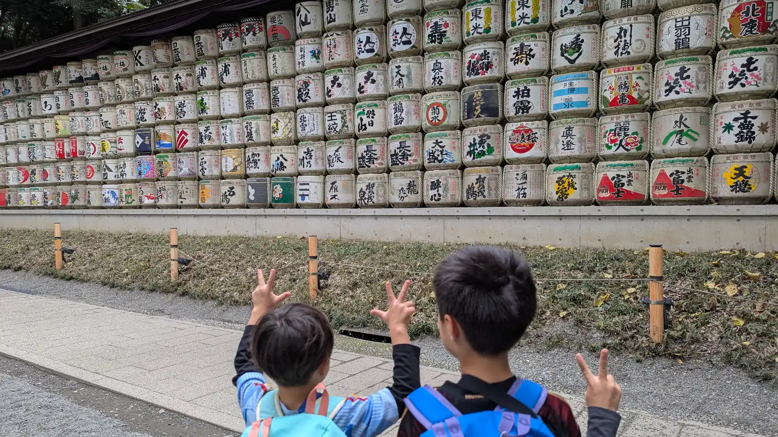 Two children standing in front of the colorful sake barrel display at Meiji Jingu Shrine in Tokyo, making playful peace signs during their visit.