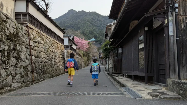 Two young boys with backpacks walk up a historic, stone-walled lane on Miyajima Island (Itsukushima), with the lush peaks of Mount Misen and a glimpse of cherry blossoms framing the background.