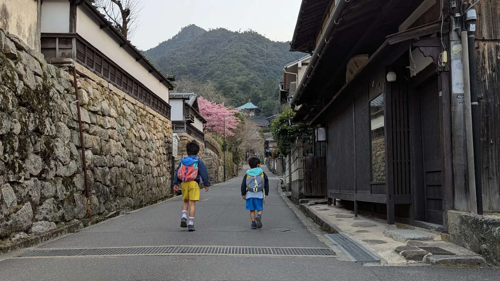 Two young boys with backpacks walk up a historic, stone-walled lane on Miyajima Island (Itsukushima), with the lush peaks of Mount Misen and a glimpse of cherry blossoms framing the background.