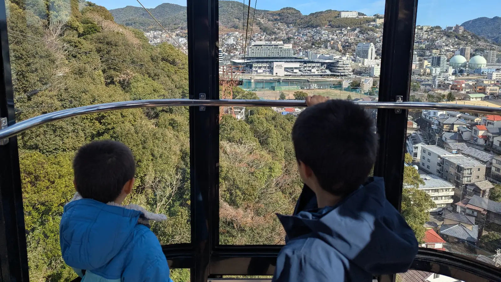 Luca & Nico riding the Mount Inasa Ropeway in Nagasaki, looking out over city rooftops and Peace Stadium with mountains in the background.