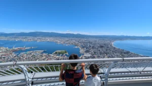 Two kids enjoy panoramic views of Hakodate city and bay from Mt. Hakodate Observatory on a sunny day.