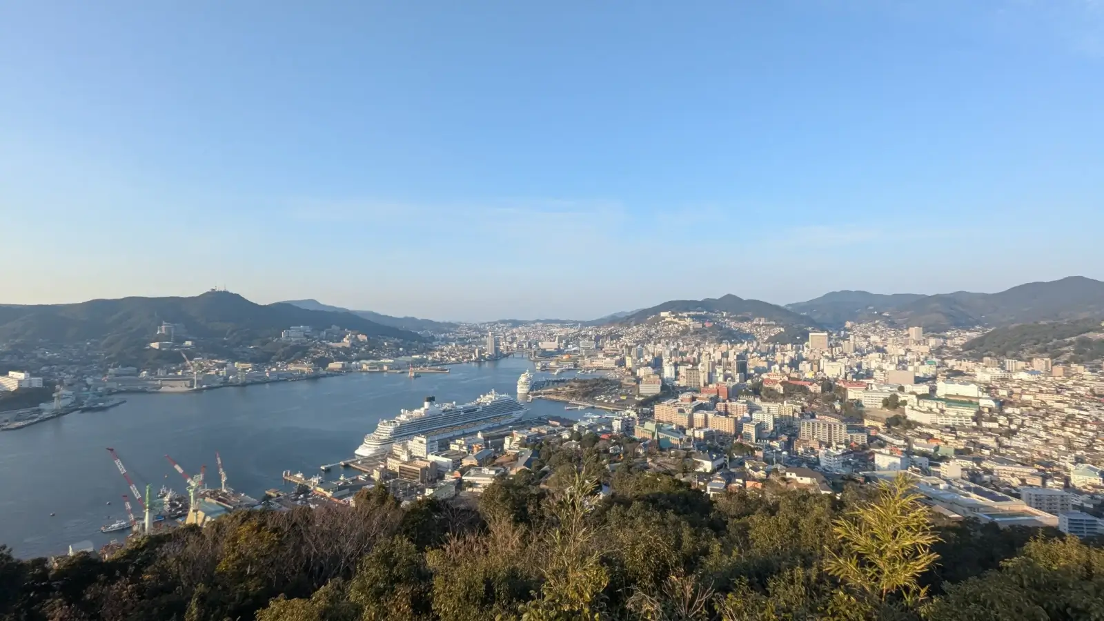 Panoramic view of Nagasaki city and harbor from the Mount Inasa observatory on a clear afternoon, capturing the full scope of the urban landscape and natural surroundings.