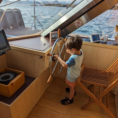 A young child in a light blue t-shirt and orange shorts stands at the helm of a wooden boat, gripping the large steering wheel while looking out at the open sea. Sunlight streams through the boat's glass windows, highlighting the calm ocean and distant islands in the background.