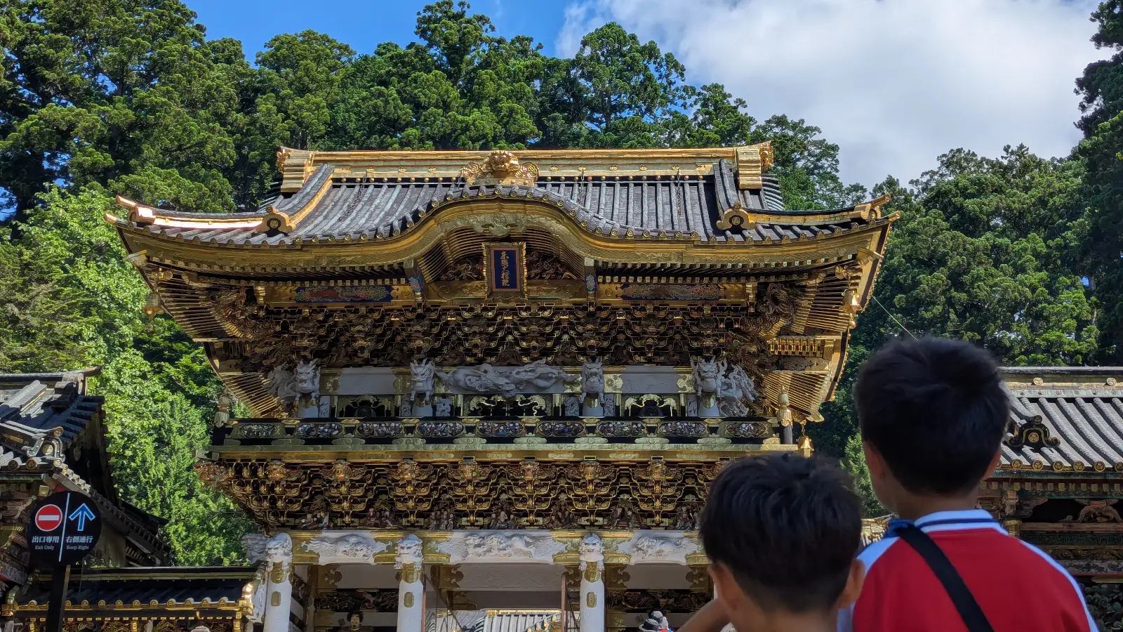 Luca & Nico looking up at the ornate Yomeimon Gate of Nikko Toshogu Shrine (日光東照宮) in Tochigi, Japan, with golden carvings surrounded by cedar trees.