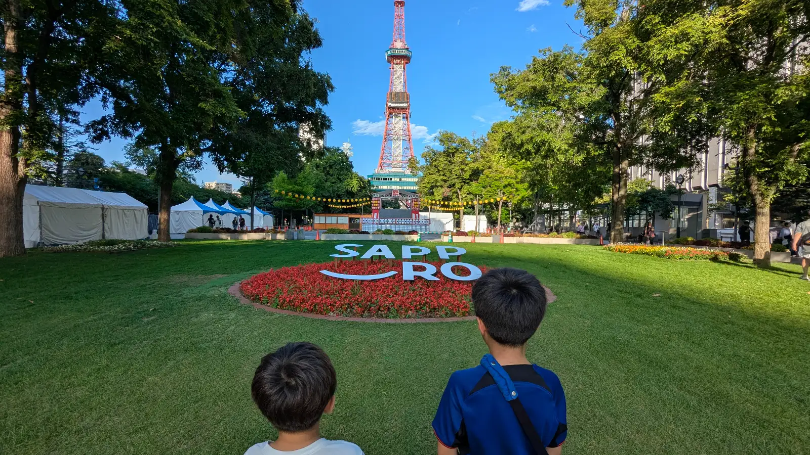 Two kids standing on the green lawn at Odori Park in Sapporo, Japan, looking toward the Sapporo TV Tower and colorful summer flowers in Hokkaido.