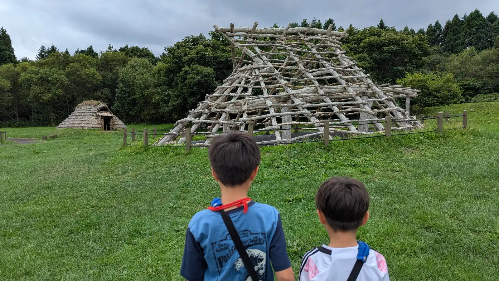 Two kids walk toward reconstructed Jomon-era pit dwellings at the Ofune Site surrounded by green grass and forest trees in Hakodate.