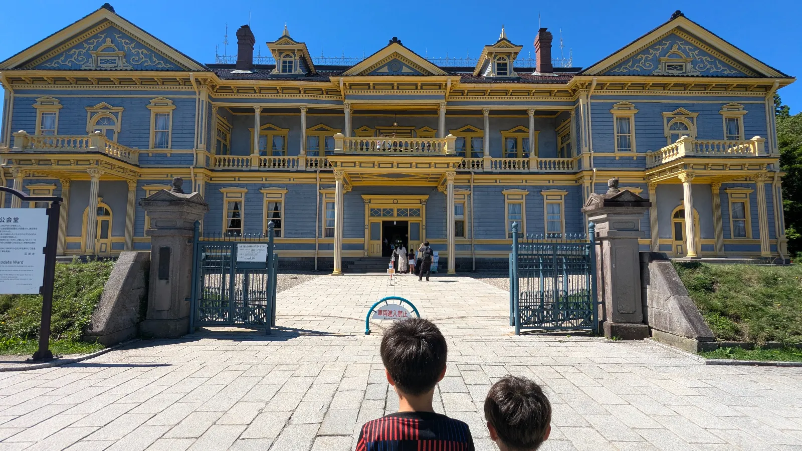 Two kids stand before the Old Public Hall of Hakodate Ward, a blue and yellow Meiji-era building with grand balconies and family visitors entering.