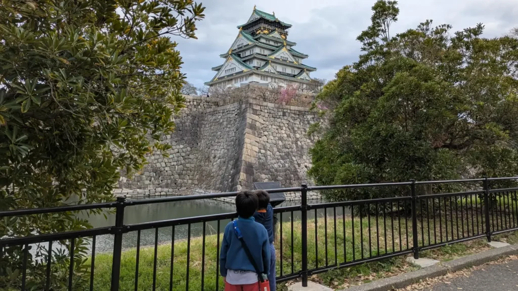 Luca & Nico gaze up at Osaka Castle from the moat path, capturing a moment of historical exploration during a family trip to Osaka, Japan.