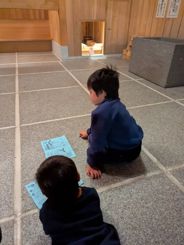 Luca and Nico study their scavenger hunt maps on the floor of the Osaka Museum of Housing & Living, surrounded by traditional wooden interiors.