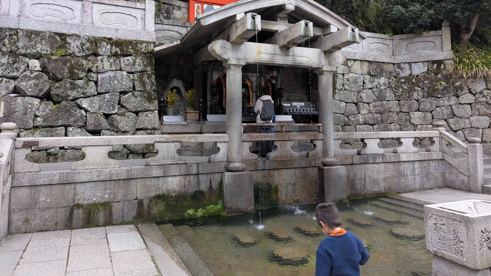 Children watch the clear streams of Otowa Waterfall flow beneath the Kiyomizu-dera Temple terrace, where visitors choose between waters for health, love, or wisdom — a memorable Kyoto family moment.