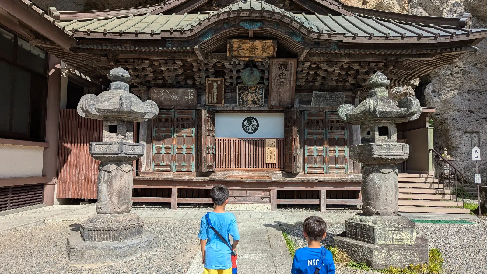Two children stand before the main wooden hall of Oya Temple in Utsunomiya, exploring its carved stone lanterns and historic architecture.