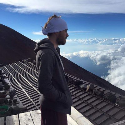 A man in a dark hoodie and white headscarf stands on a wooden platform near the summit of Mount Fuji, gazing at the sea of clouds below. The mountain's volcanic slopes and scattered rocks surround him, with a clear blue sky stretching into the horizon.