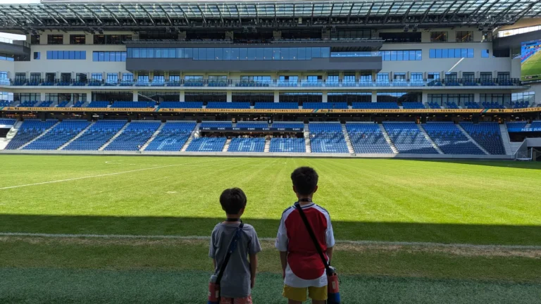 Two children stand at field level inside Peace Stadium connected by SoftBank in Nagasaki, looking out across the bright green pitch and blue stands where V-Varen Nagasaki plays home games. A peaceful family soccer adventure in Japan.