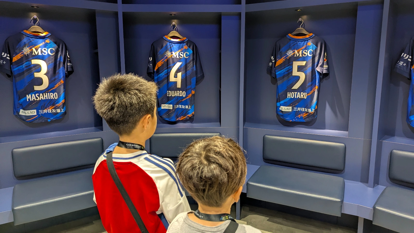 Two kids look up at the V-Varen Nagasaki players’ jerseys inside the team locker room at Peace Stadium connected by SoftBank, part of a family tour showing children’s perspective of Japanese soccer culture.