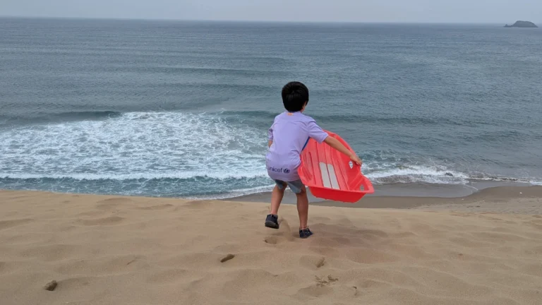 Boy holding a sled at the Tottori Sand Dunes with the Sea of Japan in view, ready for sandboarding adventure.