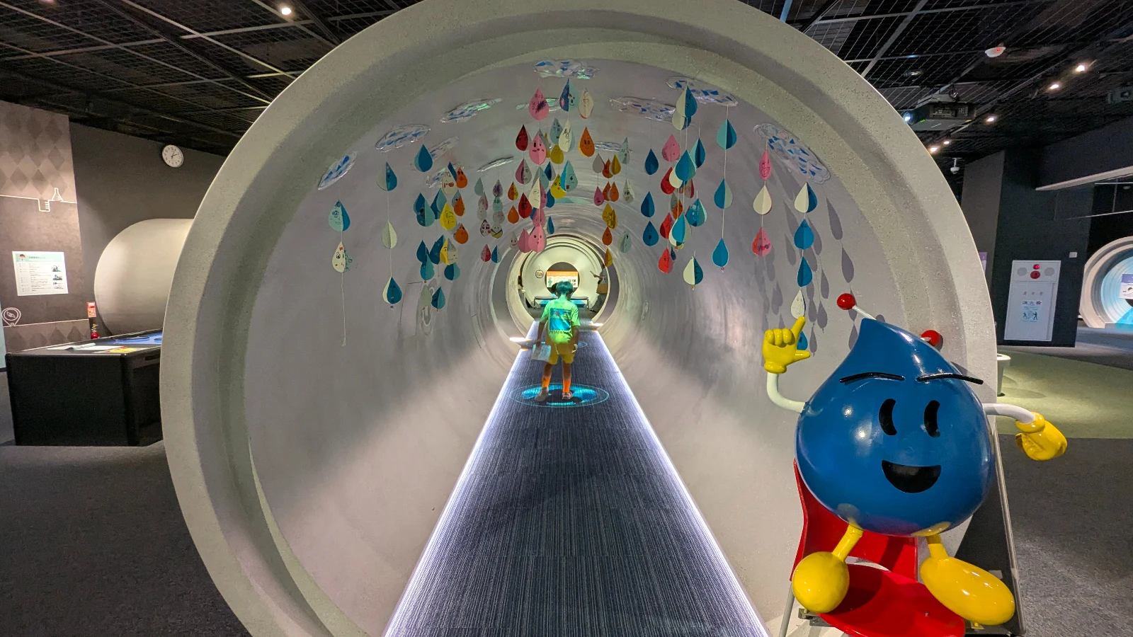 Child exploring the large pipe tunnel exhibit at the Sapporo City Sewage Science Museum with colorful raindrop decorations and a blue sewer mascot.