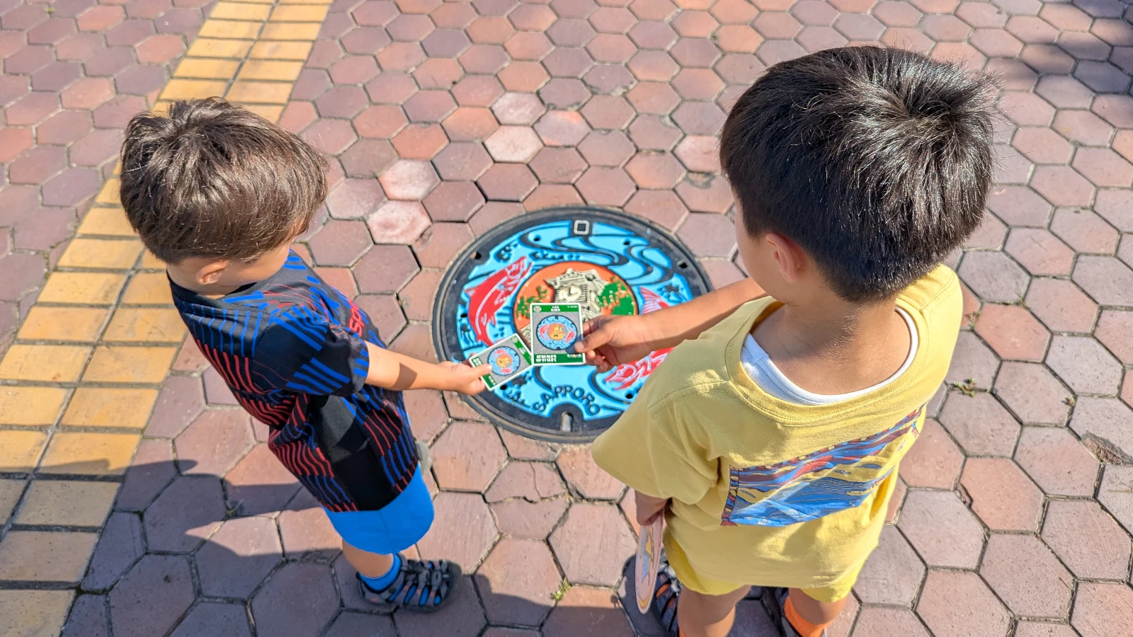 Two boys holding collectible manhole cards above a colorful Sapporo manhole cover outside the Sapporo City Sewage Science Museum in Japan.