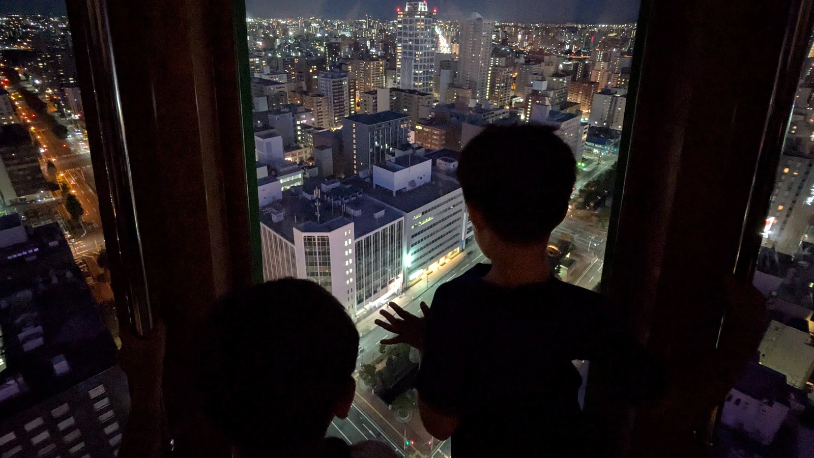 Two children gaze out from the Sapporo TV Tower observation deck at the city lights below, enjoying the panoramic night view of Sapporo’s skyline.