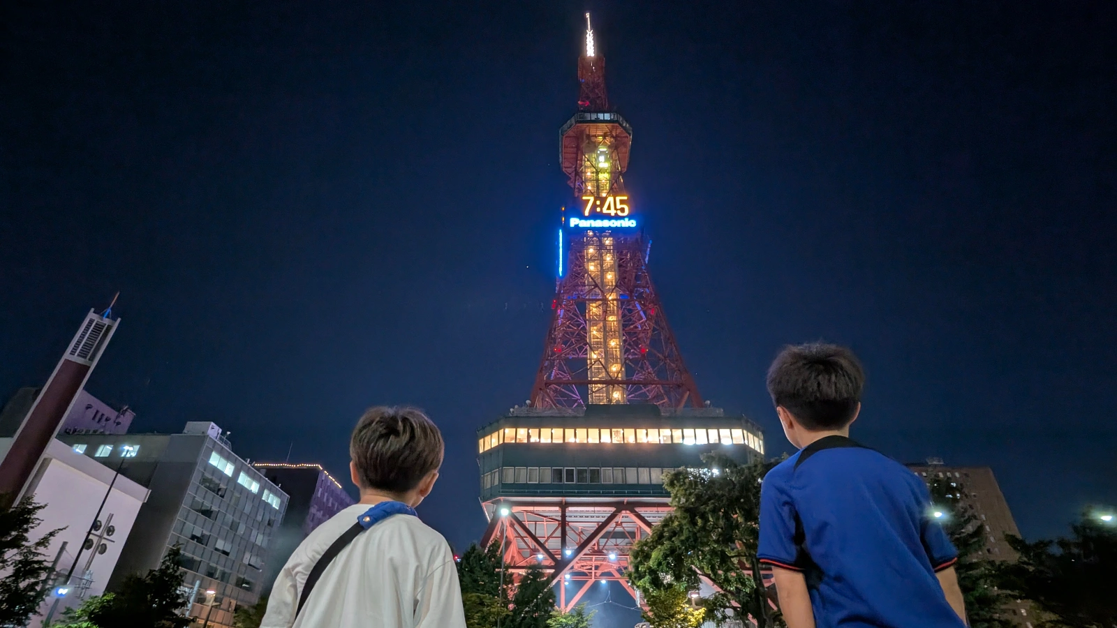 Two children look up at the illuminated Sapporo TV Tower at night in Odori Park, capturing the tower’s glowing clock and observation deck during an evening family visit.