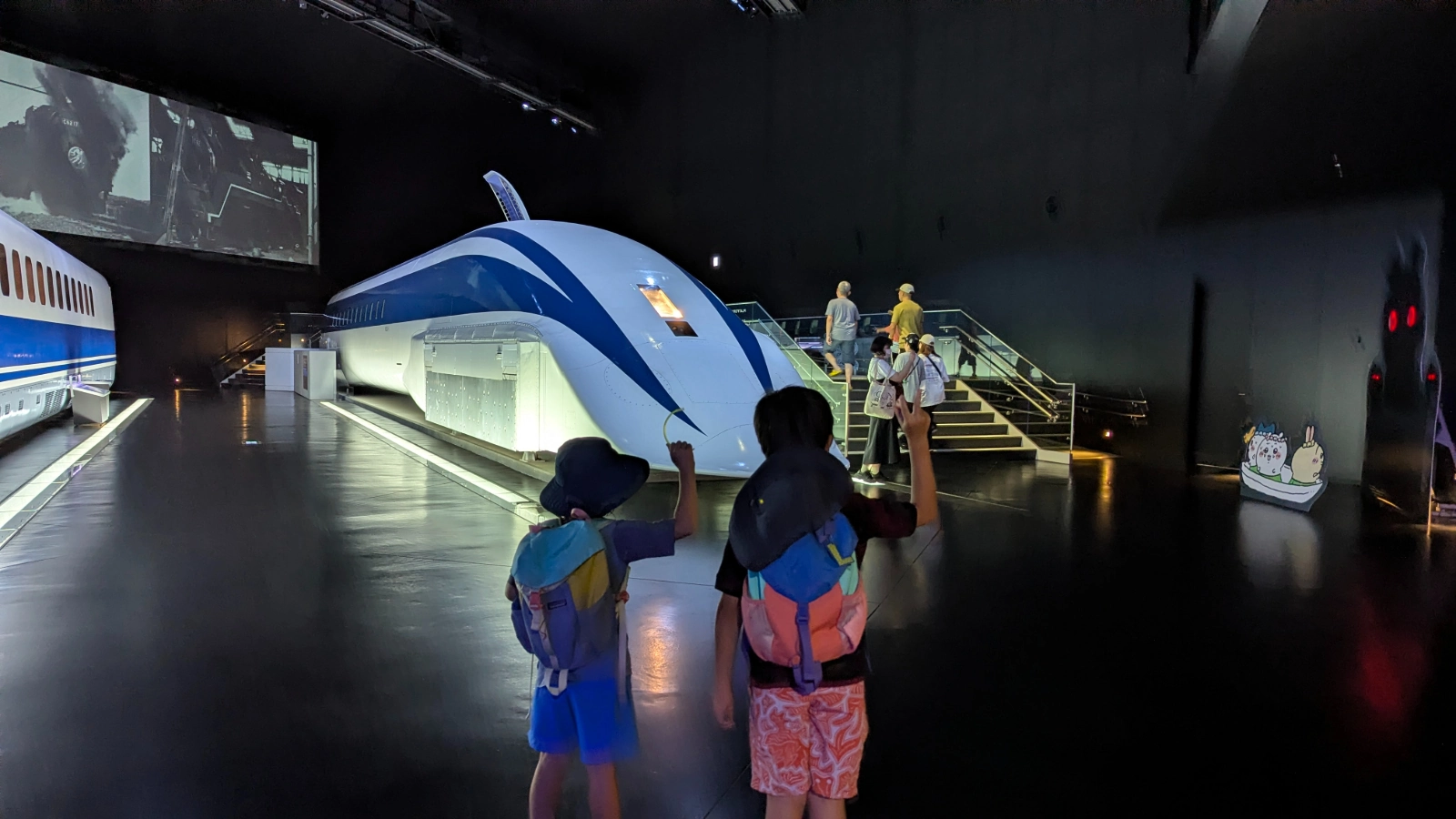 Luca and Nico wave at a maglev test car in a darkened hall at SCMaglev and Railway Park in Nagoya.