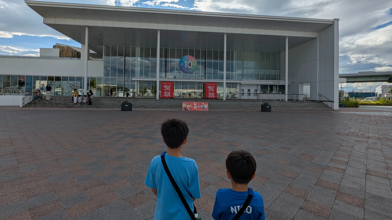 Two children stand in front of the modern glass entrance of Sendai Umino-Mori Aquarium on a sunny afternoon in Sendai, Japan.