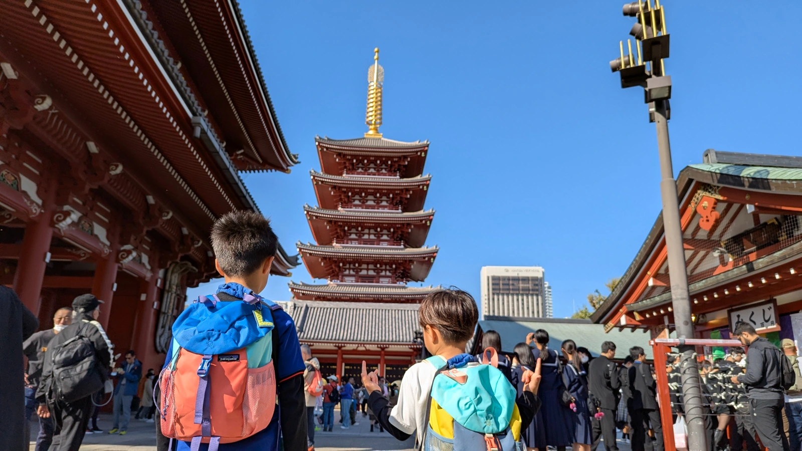 Two kids looking up at the five-story pagoda at Senso-ji Temple in Asakusa, Tokyo, with bright blue skies and temple buildings around them.