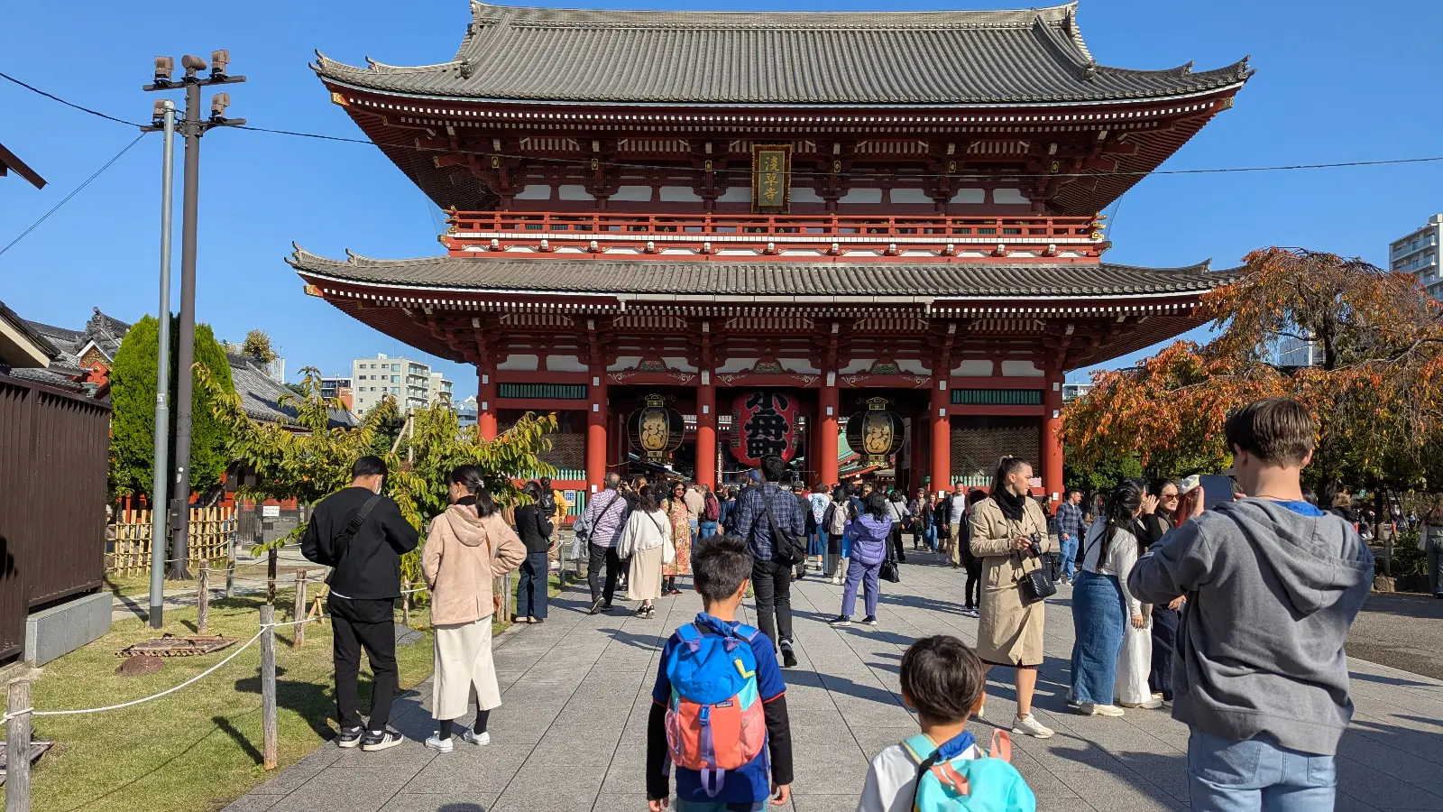 Two boys with backpacks approaching the Hozomon Gate at Senso-ji Temple in Asakusa, Tokyo, on a sunny day with many visitors around.