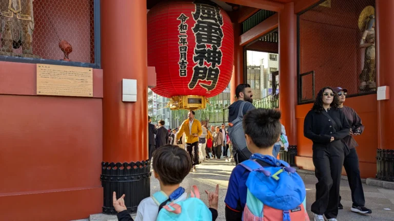 Two boys standing beneath the giant red lantern of the Kaminarimon Gate at Senso-ji Temple in Asakusa, Tokyo, watching crowds pass through.