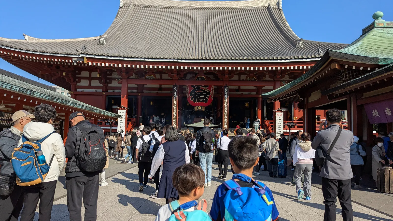 Two kids with backpacks walking toward the main hall of Senso-ji Temple in Asakusa, Tokyo, surrounded by visitors under a bright blue sky.