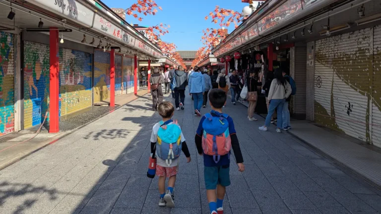 Two kids walking down Nakamise Shopping Street toward Senso-ji Temple in Asakusa, Tokyo, lined with colorful shop shutters and autumn decorations.
