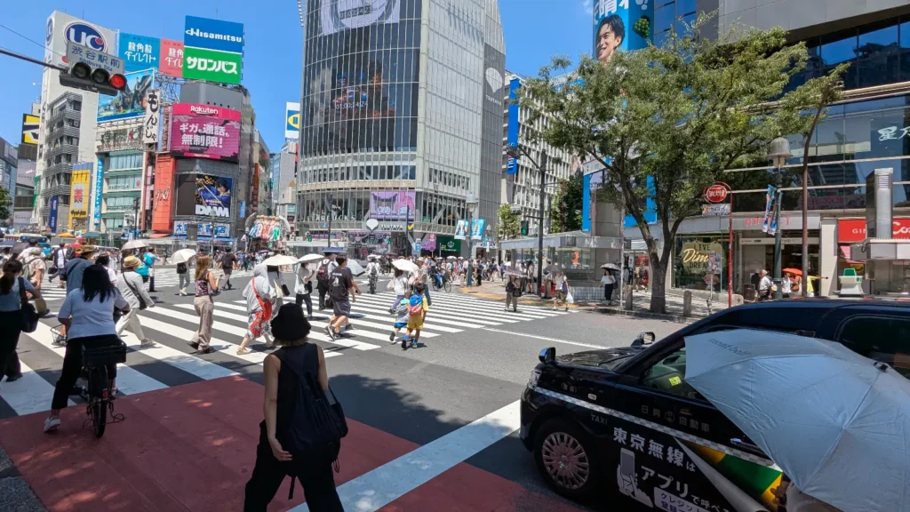 Luca & Nico with crowds of people crossing Shibuya Scramble Crossing in Tokyo during the day, with neon billboards and observation deck buildings in the background.