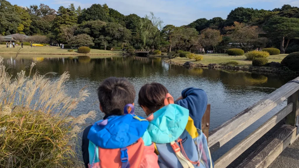Kids peering over a bridge railing at the pond in Shinjuku Gyoen National Garden, surrounded by autumn trees.