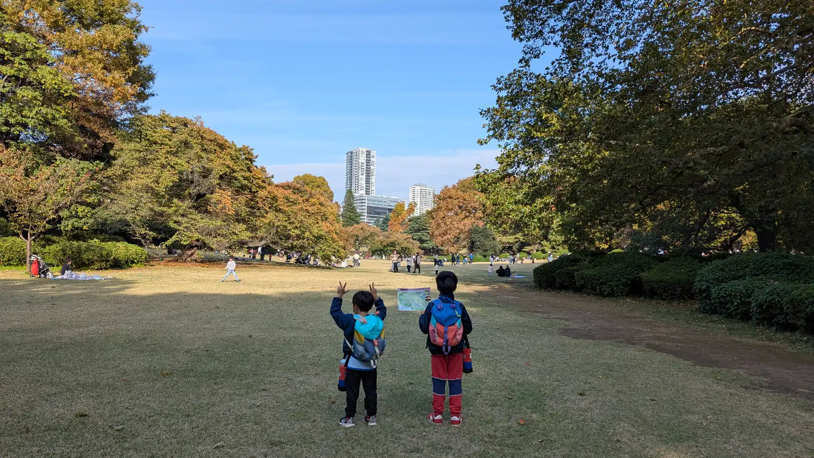Kids standing on the open lawn at Shinjuku Gyoen National Garden with autumn trees and Tokyo skyline in the background.